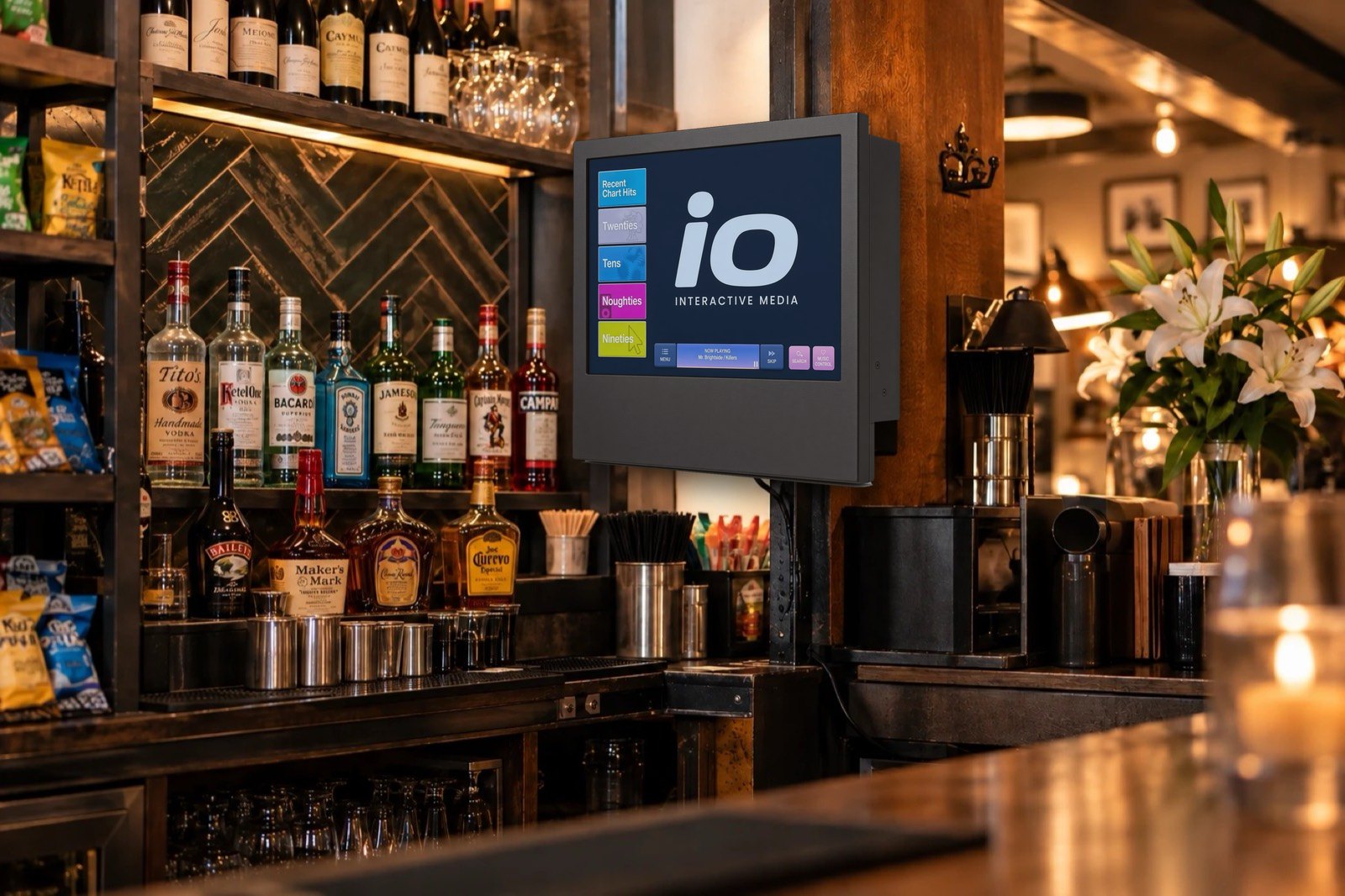 Wall-mounted iO Interactive Media touchscreen behind a bar showing the music selection screen
