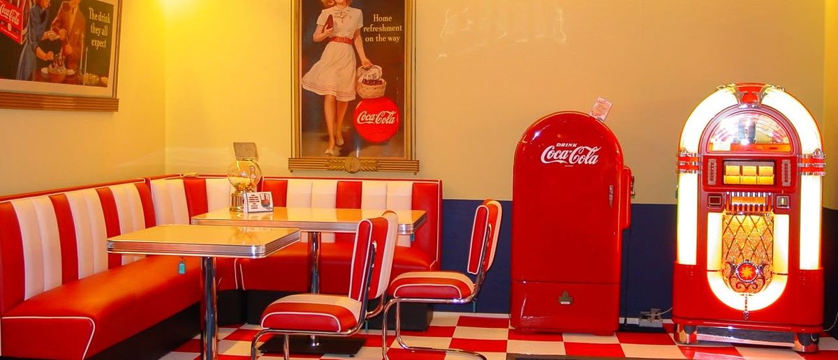 Retro diner-style room with red booths and a glowing jukebox beside a vintage drinks fridge