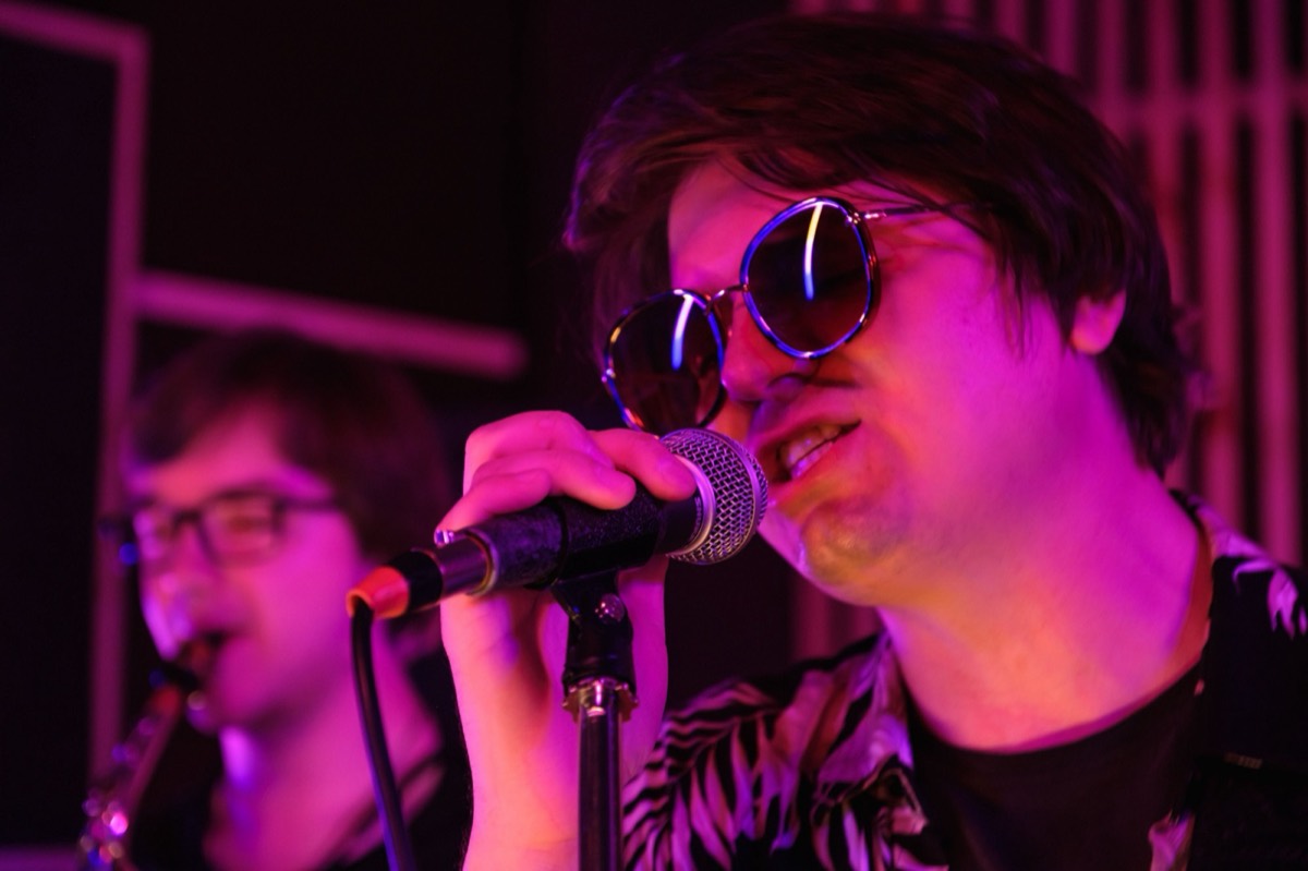 Singer performing in a colourful, atmospheric room with sunglasses and stage lighting